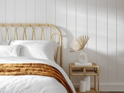 Un dormitorio con cabecero de ratán, ropa de cama blanca y una manta decorativa de color marrón anaranjado. Una mesita de noche pequeña sostiene un jarrón decorativo con hojas secas y otros elementos pequeños. Se ven paredes con paneles blancos que capturan la esencia de la fotografía inmobiliaria a la perfección.