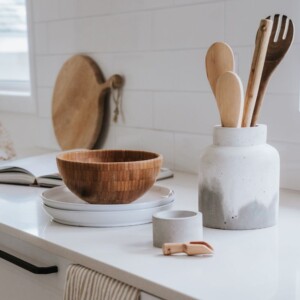 Una encimera de cocina con un cuenco de madera sobre platos blancos, un tarro de cerámica con utensilios de madera y un pequeño recipiente de cerámica al lado. Una tabla de cortar y un libro abierto se encuentran en el fondo, perfectos para una publicación inmobiliaria o una sesión fotográfica con estilo.