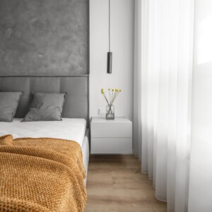 Un dormitorio moderno con un cabecero tapizado en gris, almohadas grises, una manta naranja, una mesita de noche blanca y un jarrón con flores. Unas cortinas blancas transparentes cubren una ventana, lo que permite que entre la luz natural, perfecta para fotografía inmobiliaria.